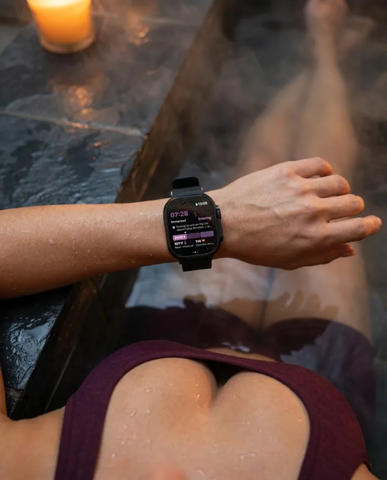 First-person view from a steaming hot tub: the wearer's own wrist resting on the stone rim, Apple Watch showing a spa session 7:28 immersed, Zone 1, water 101°F measured, with a note that resting an arm on the rim doesn't stop the clock.